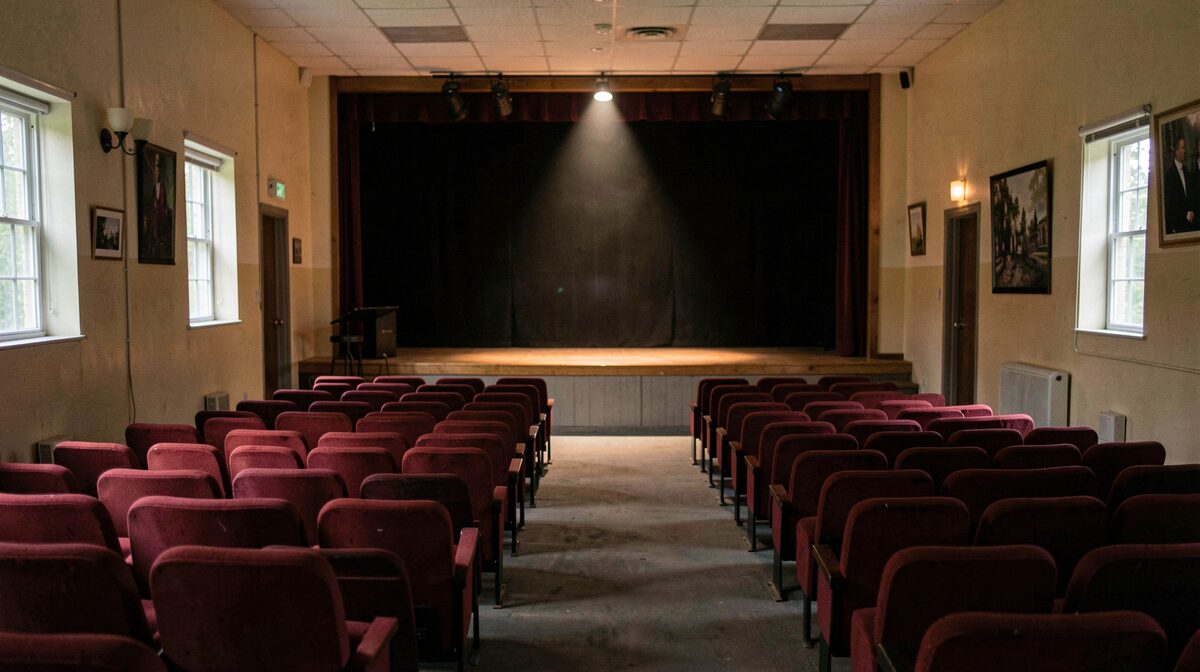 Interno di una piccola sala spettacoli di provincia con file di sedie rosse, luci spente e un solo riflettore sul palco vuoto, inquadrato dal centro della platea
